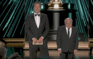 Oscars 2024 gif. Arnold Schwarzenegger clutches an award envelope in front of him while shifting side to side. Danny Devito looks around him in awe and says, "Very nice."