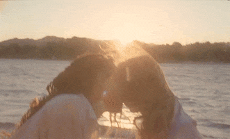 Music video gif. From the video for Chance, Hayley Kiyoko and Alexandra Shipp kiss in the glow of a sunset while gentle waves churn in the landscape behind them.