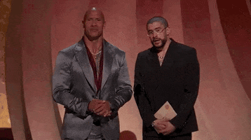 Oscars 2024 GIF. Bad Bunny and The Rock are presenting the award for Best International Feature. Both men have their hands clapsed in front of them and Bad Bunny is holding the envelope. He speaks in Spanish, which is translated to English subtitles at the bottom, and he says, "Cinema is a universal language that speaks to the common threads that bind us together." 