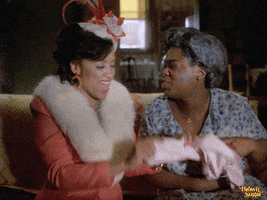 TV gif. Two Black women, seated on a couch together, dressed in vintage wear, throw their heads back, cackling with joy.