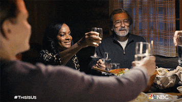 TV gif. Table full of characters on This is Us including Justin Hartley as Kevin and Griffin Dunne as Nick Pearson lean forward to clink their different glasses together to make a toast.