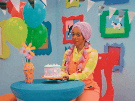 TV gif. Character in Happy Place wearing a lavender pastel wig, bright yellow checkered shirt, and coral-colored overalls sits at a table holding up a birthday cake in a brightly colored cartoonish set. She smiles sweetly and says, "Happy birthday bestie."