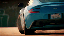 Video gif. Blue Aston Martin spins donuts over a dusty road, kicking up a cloud in its wake.