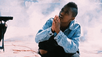 Music Video gif. Syd the Kyd in an The Internet music video kneels on the ground amongst smoke and prays up to the sky.
