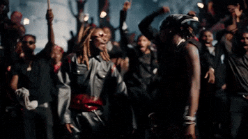 Music video gif. Fireboy DML and Asake are in their music video dancing as a crowd behind them also gets hype, jumping to the beat.