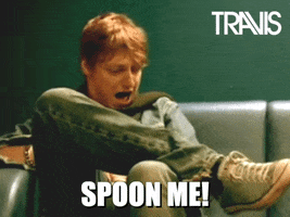 Celebrity gif. Sitting cross-legged on a couch, Dougie Payne exclaims with surprise and a spoon floats up from his lap up to cover his eye. Text, "Spoon me!"