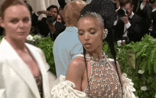Met Gala 2024 gif. Stella McCartney approaching FKA Twigs from behind and throwing an arm over her shoulder an leaning in, posing for the cameras, FKA Twigs in a sheer beaded ensemble with loopy sweater-like boa, Stella in a white suit jacket, both by Stella McCartney.