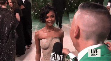 Met Gala 2024 gif. Tyla gives a sassy eye roll with a flirty smile during an interview. The top of her sand-colored dress is sculpted to look like formed wet sand that is caked to her body revealing the shape and details of her torso. 
