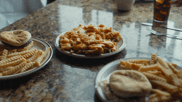 Movie gif. In a scene from An Evening with Beverly Luff Linn, a hand reaches toward a plate of gooey chili cheese fries, taking a greedy handful.
