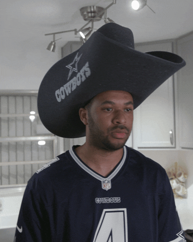 Video gif. Man wearing a Dallas Cowboys jersey and an oversized foam cowboy hat shakes his head in disbelief as he looks off with a focused expression.