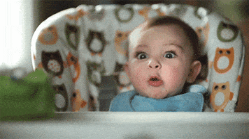 Video gif. Sitting in a high chair, baby appears excited as his eyes widen and spit bubbles form in his pursed lips.