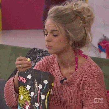 Video gif. A young woman with a messy bun digs into a bag of popcorn, then turns with surprised eyes and smiles.
