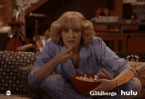 TV gif. Wendi McClendon Covey as Beverly in The Goldbergs, sits on a couch hunched over and eating out of a red bowl of popcorn. She leans forward with wide eyes glued to the screen ahead of her. 