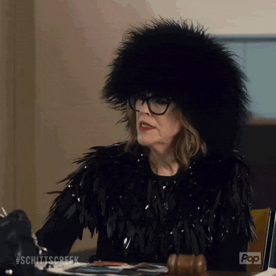 Schitt's Creek gif. Catherine O'Hara as Moira Rose is sitting at a judge's table. She sits upright and claps her hands daintily near her face before jotting down a score.