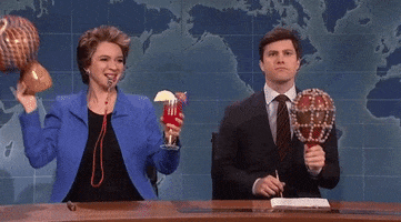 SNL gif. Holding a cocktail, shaking a beaded maraca, and blowing a coach's whistle, Maya Rudolph sits next to Colin Jost on Weekend Update, who shyly shakes a maraca.