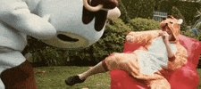 Music video gif. From a Daydreamers video, a person in a cow mascot costume headbutts a guy in a giraffe onesie, who falls over onto the grass off of his red inflatable chair.