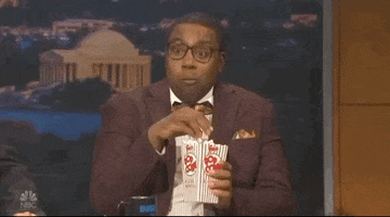 SNL gif. Kenan Thompson wears glasses and a brown suit with a bow tie as he munches on popcorn and nods earnestly.