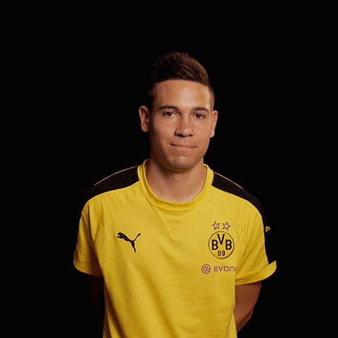 Sports gif. Raphael Guerreiro in his Borussia Dortmund jersey, smiles at us and raises his hand like he's showing the height of something. 
