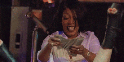 Celebrity gif. K Michelle smiles as she drops dollar bills between a woman's legs that jiggle in the air doing a split in front of her. 