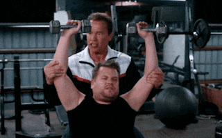 Late Night Show gif. James Cordon holds two normal sized exercise weights over his head as Arnold Schwarzenegger spots him, holding his arms up. James Corden drops the weights and pants exaggeratedly like he’s overly exhausted by the pretty small weights.