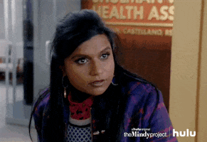 TV gif. Mindy Kaling as Mindy Lahiri from The Mindy Project enters her workplace and genuinely asks, "What day is it?" A bunch of her coworkers stand at white tabletops surrounding a decorative Christmas tree and immediately stop what they're doing to turn and stare at her with wide eyes.