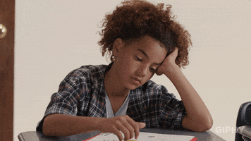 Video gif. Bored child rests their head on their hand as they doodle on a sheet of paper at a school desk, looking up at the front of class with very little interest.