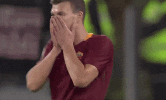 Sports gif. Edin Džeko winces and buries his face in his hands, then bends over on a soccer field with other players.