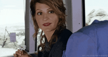 Movie gif. Nia Vardalos as Georgia in My Life in Ruins sits on a bus and looks over her shoulder while holding a white flower in her hand. She gives an exaggerated, cheeky wink at someone.
