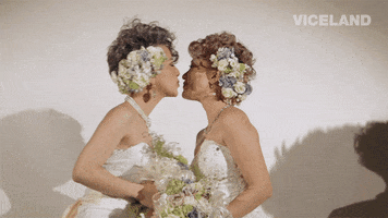 Reality TV gif. Two women on Gaycation hold bouquets and wear wedding dresses as they kiss each other happily. 