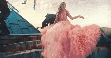 Music video gif. Taylor Swift sits in an elaborate ruffled pink dress and sings her song Me, while Brendon Urie of Panic at the Disco descends from the sky using an umbrella behind her. Two dancers twirl black umbrellas on each side of him.