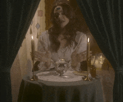 Video gif. Woman dressed as a fortune teller sits at a circular table with candles and tarot cards. She waves her hands over a crystal ball and text appears, "I seeee... A crying cat in your future." She looks up at us with insincere pity. 