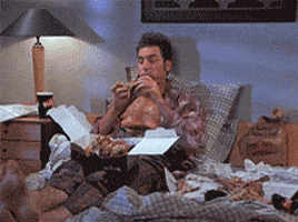 Seinfeld gif. Michael Richards as Kramer sitting in bed, tapping his feet, and contentedly chowing down on a box of fried chicken.