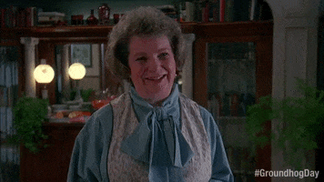 Movie gif. Angela Paton as Mrs. Lancaster in "Groundhog Day," wears a turquoise shirt with a bow and a vest in front of a bookcase, cocks her head to the side as she smiles.