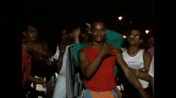 Video gif. Man in a vibrant red tank top confidently vogues in a crowd, getting down low as another man in a green shirt vogues on top of his head.