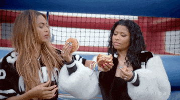 Celebrity gif. Beyoncé and Nicki Minaj wear deluxe sports apparel as they intertwine arms and take big bites of burgers. 