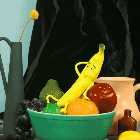 Stop-motion gif. Zooming in on a tabletop holding a bowl of fruit to show a banana reclining flirtatiously and winking.