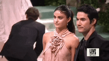 Met Gala 2024 gif. Slow motion closeup of the top of Camila Mendes and Joseph Altuzarra's outfits. Mendes is wearing a dusty rose halter dress with a rosebud detailing where the straps meet at the center of her chest. The dress has delicate flowing vertical panels that hang straight. Her hair is parted at the top of her forehead and swept back in a ponytail. Altuzarra is wearing a black shiny suit jacket and shirt.