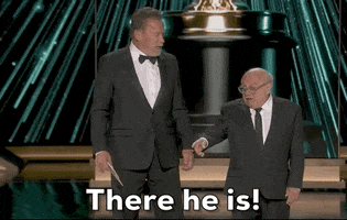 Oscars 2024 gif. Arnold Schwarzenegger looks down at Danny Devito, who points into the crowd and says, "There he is!"