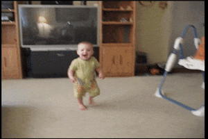 Video gif. Baby happily runs toward us in a living room, then a cat jumps in the frame to attack and knocks the baby to the carpet, where he lays crying.