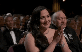 Oscars 2024 GIF. Lily Gladstone smiles towards the stage while she softly claps. 