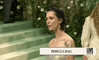 Met Gala 2024 gif. Rebecca Hall wearing Danielle Frankel and watercolor flower body art, poses for the cameras then snaps out of it with a confident chuckle and moves on.