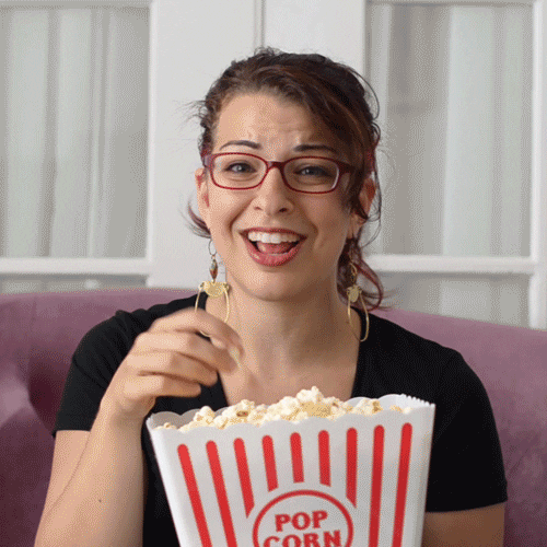 Video gif. Woman is eating from a big carton of popcorn and she looks engrossed as she munches and watches her show.