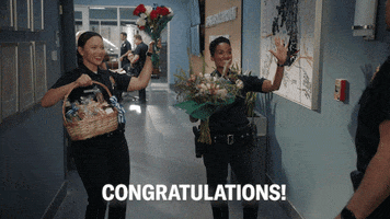 TV gif. On The Rookie, an excited Mekia Cox as Nyla offers a bouquet while Melissa O’Neil as Lucy offers a gift basket and roses to a coworker. Text, “Congratulations!”