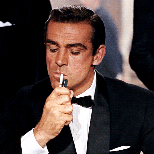 Movie gif. Sean Connery as James Bond. He has a cigarette in his mouth and he lights it, puffing and looking up.