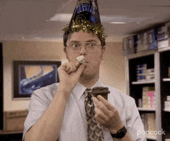 TV gif. Clip of Rainn Wilson as Dwight in "The Office" wearing an "Over the Hill" party hat and blowing on a noisemaker, giving a somewhat unsettling and overenthusiastic smile while holding a chocolate cupcake with a single unlit candle. Suddenly, Ed Helms as Andy bursts into frame, peeling around an open door with a big cheesy smile on his face. 