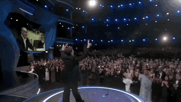 Oscars 2024 gif. Robert Downey Jr walks onstage and faces a vast standing ovation. His arms are raised as he gives two peace signs to the crowd. 