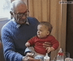 Video gif. Grandbaby sits on his grandfather’s lap as the grandpa offers something delicious to the baby on the tip of his finger. The baby excitedly accepts the treat, and the grandfather reacts in amusement before the baby bites down on the grandfather’s finger, making him cry out in shock and pain as the baby looks around in confusion.