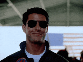 Movie gif. Tom Cruise as Maverick in Top Gun smiles warmly as he puts sunglasses on. 