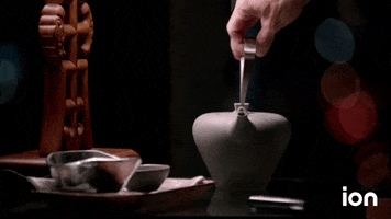 Video gif. Hand pours clear liquid out of a sleek gray teapot into a small gray cup. An intricate piece of smooth woodwork rests behind the cup, and the background is dark with hazy glints of red and blue lights.