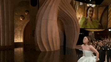Oscars 2024 GIF. Emma Stone wins Best Actress. She gets up on stage and emotionally beckons to the back of her dress to signal that it's broken. She turns around to show the presenter. 